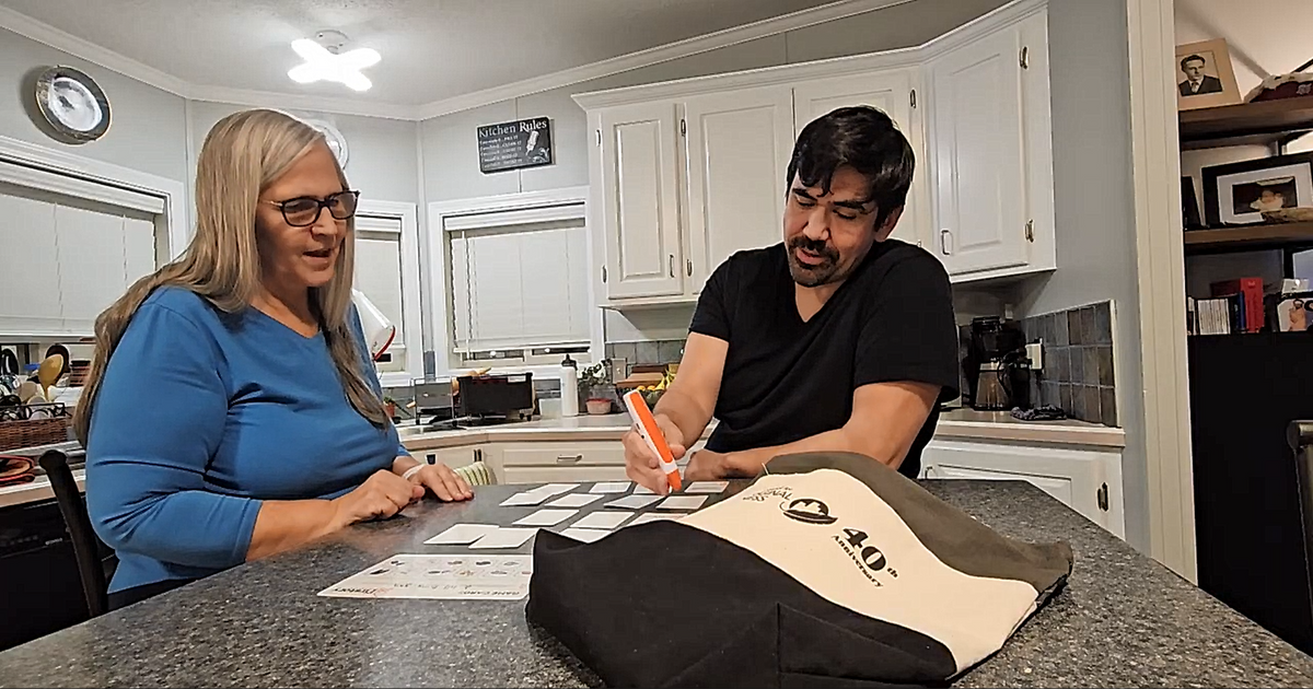 A man and a woman sitting at a kitchen counter, looking at printed language cards while using a language pen. A canvas tote bag with a black and cream logo sits on the counter in the foreground.